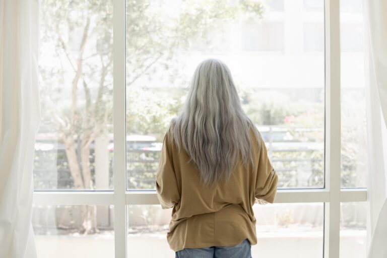 Donna con desiderio responsivo di spalle davanti a una finestra, avvolta in un maglione, guarda fuori in una luce chiara